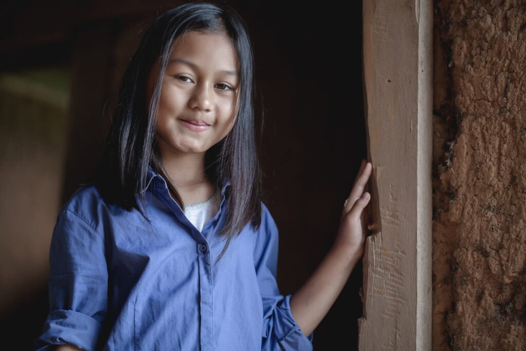 Dziewczynka w niebieskiej koszuli stojąca przy drewnianej ścianie, symbolizująca rozwój i edukację w ramach funduszy europejskich, związana z projektami wspierającymi dzieci i młodzież.